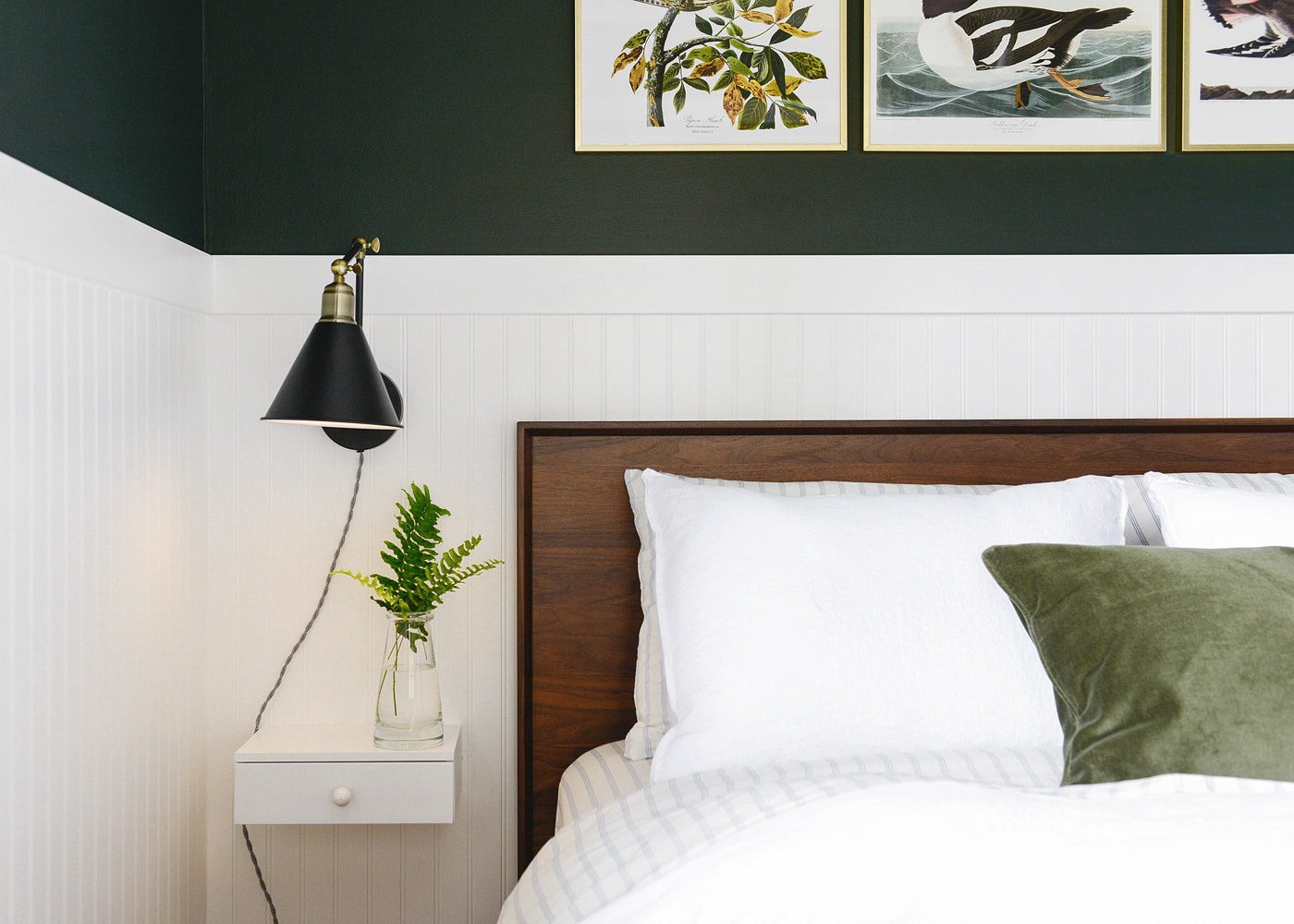 A walnut platform bed with white bedding, black bedside sconce and dark green walls above white beadboard | via Yellow Brick Home