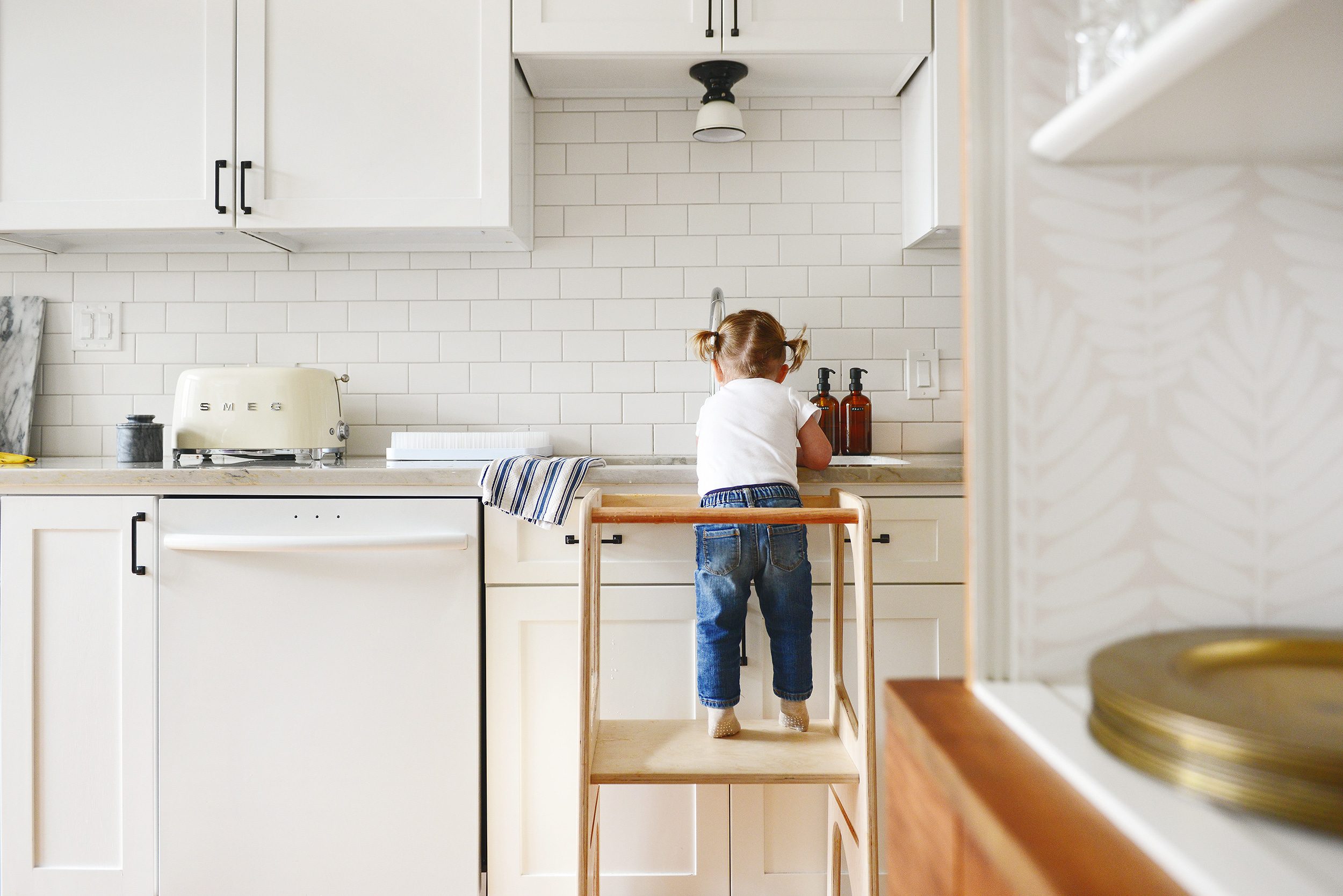 A toddler uses an activity stand to wash her hands in the sink of a classically styled all-white kitchen // updates on our most popular projects via Yellow Brick Home