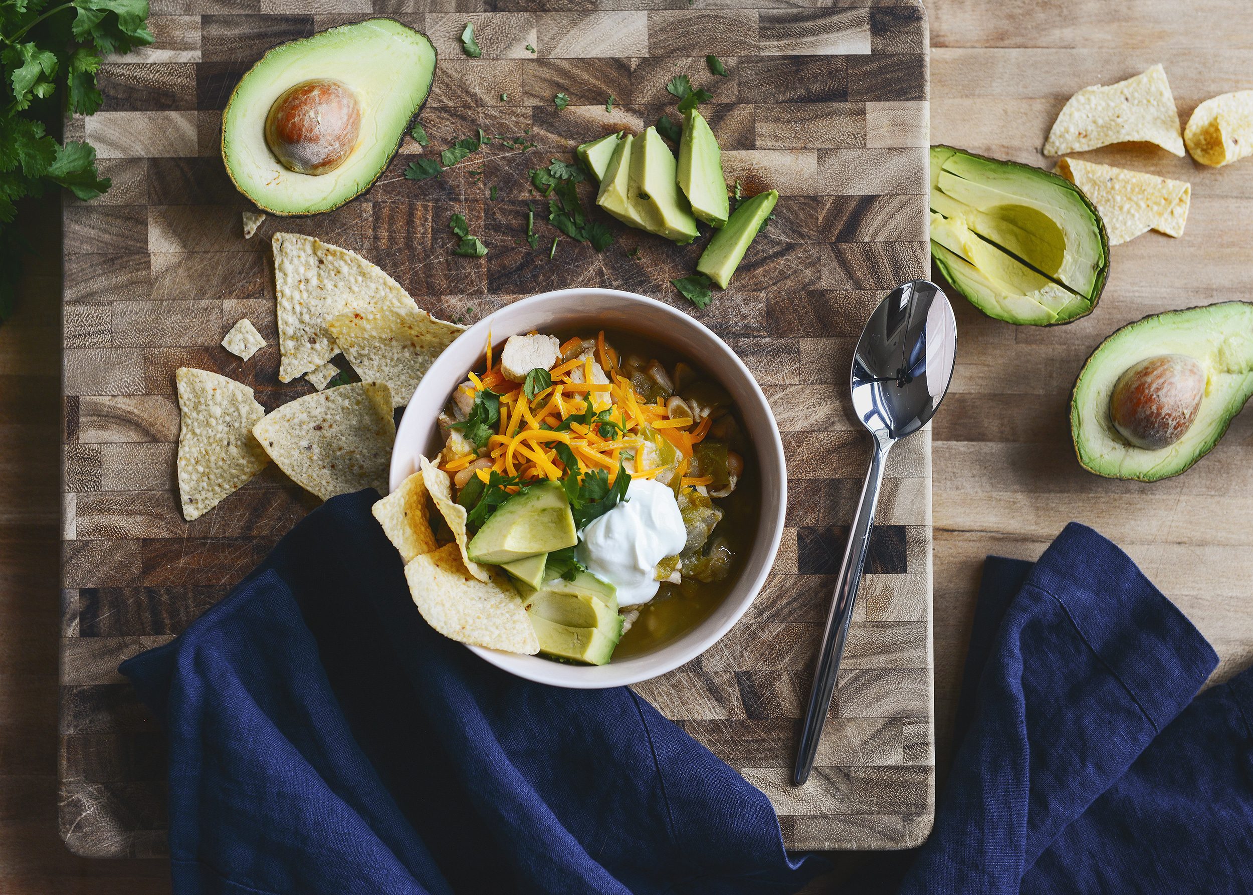 A bowl of warm chili with avocado and navy linen napkins | Our White Chicken Chili! Full of tomatillos and chunky chicken, we top it with sliced avocado and heaps of cilantro. via Yellow Brick Home