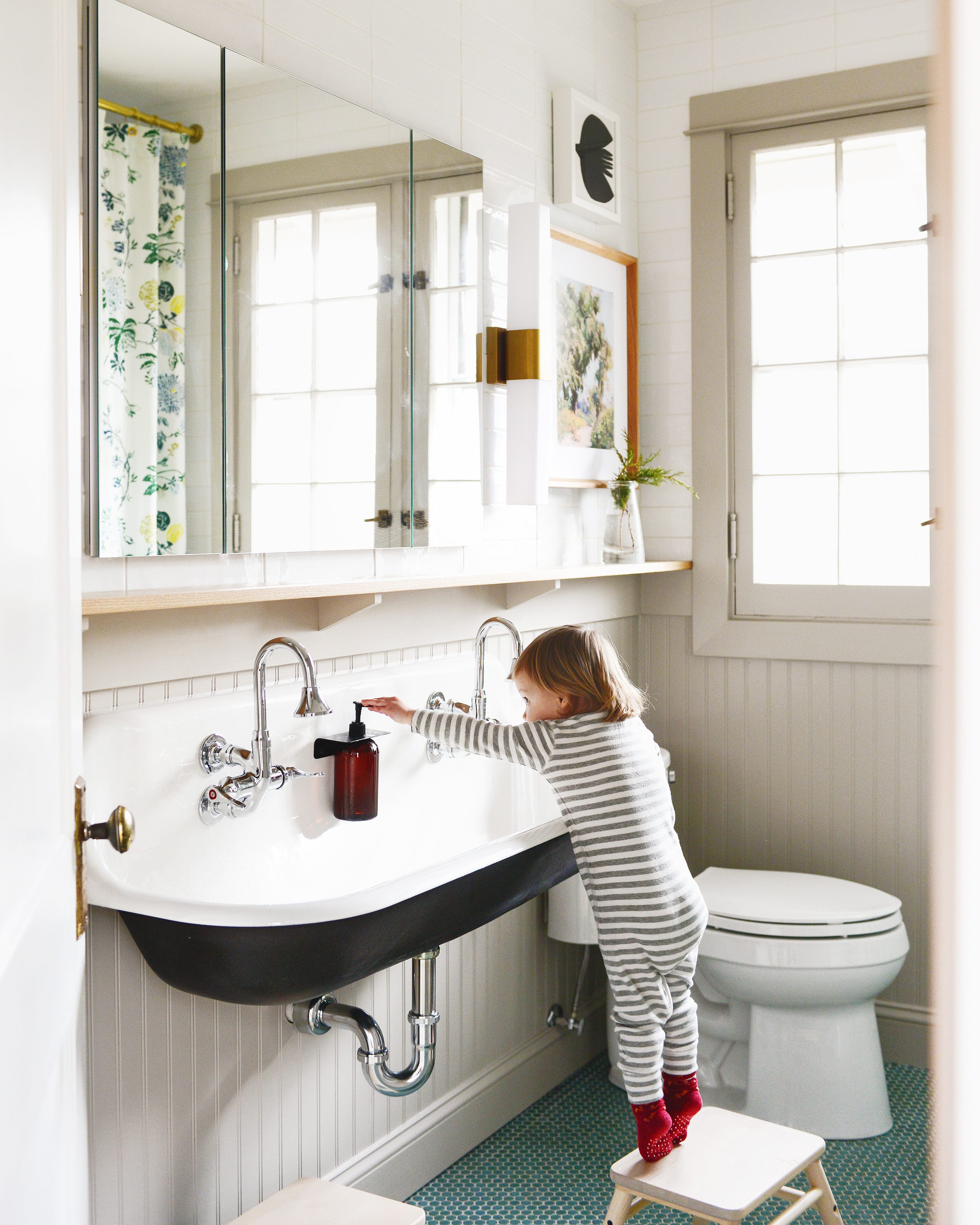 Lucy stands on a step stool in a neutral bathroom, reaching for the soap pump. The walls are beadboard and the floor is green penny tile. via Yellow Brick Home