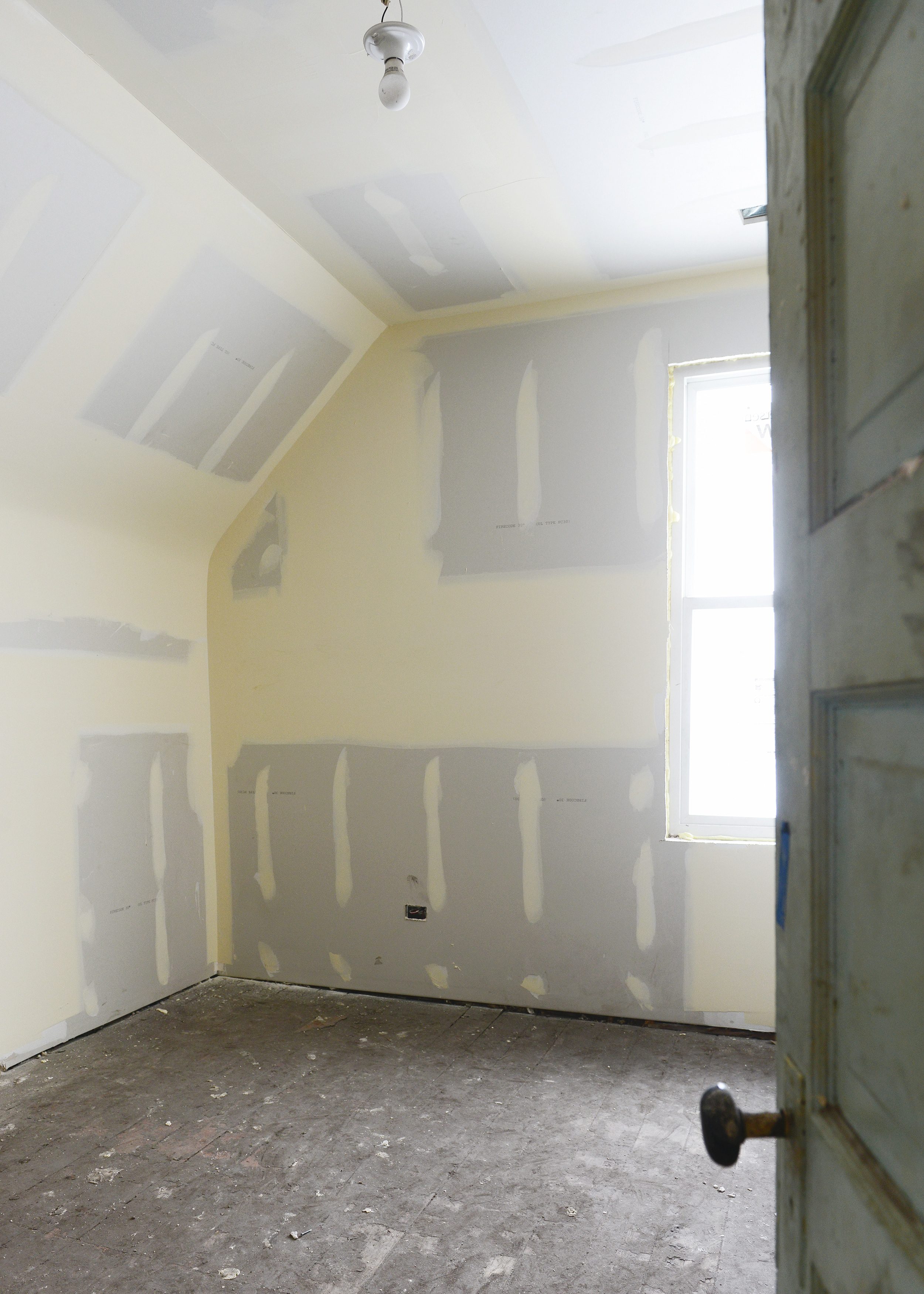 A vintage door opens into a freshly drywalled bedroom with a sloped ceiling