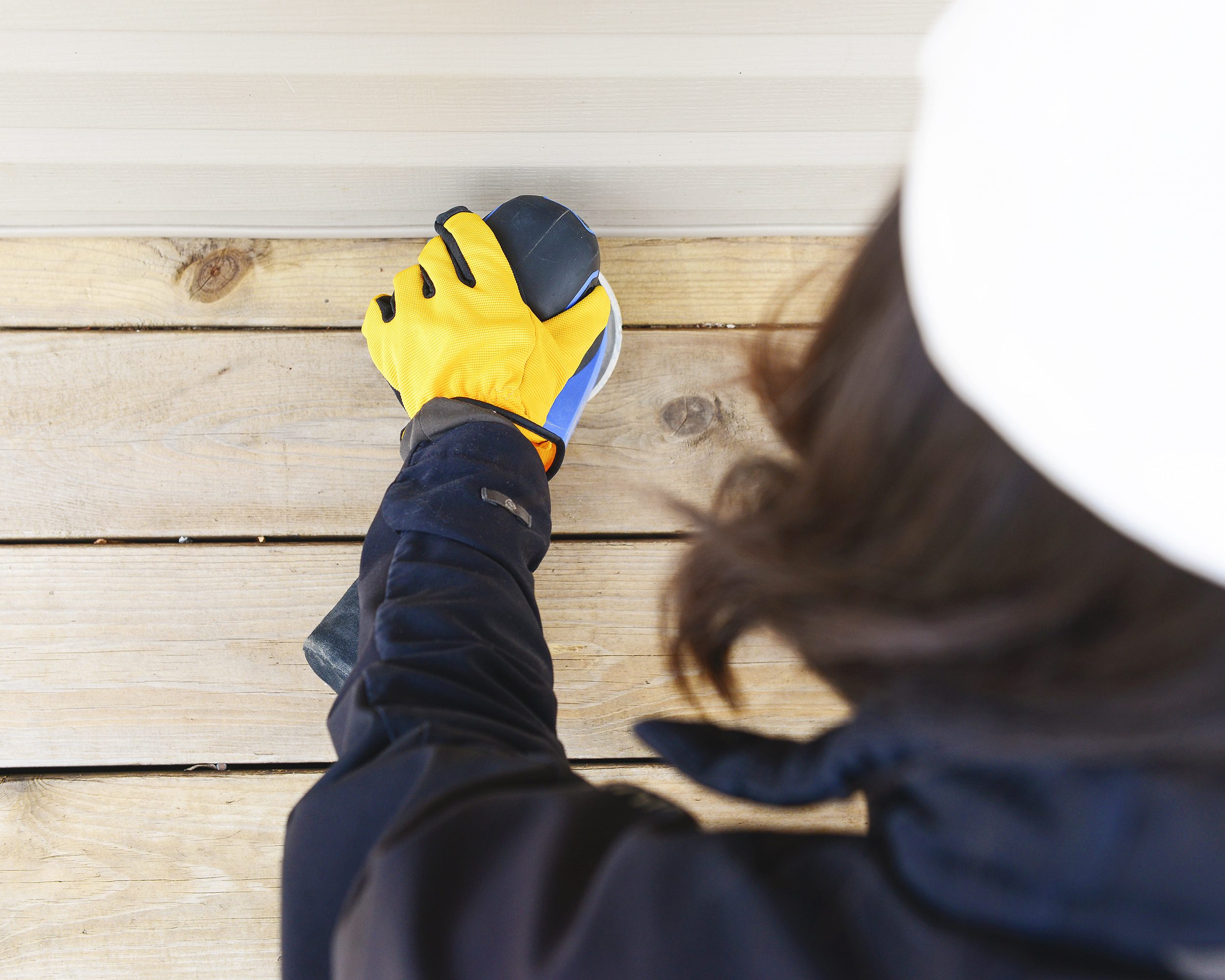 A woman in a white baseball cap and yellow work gloves sands a deck with a blue palm sander // via Yellow Brick Home