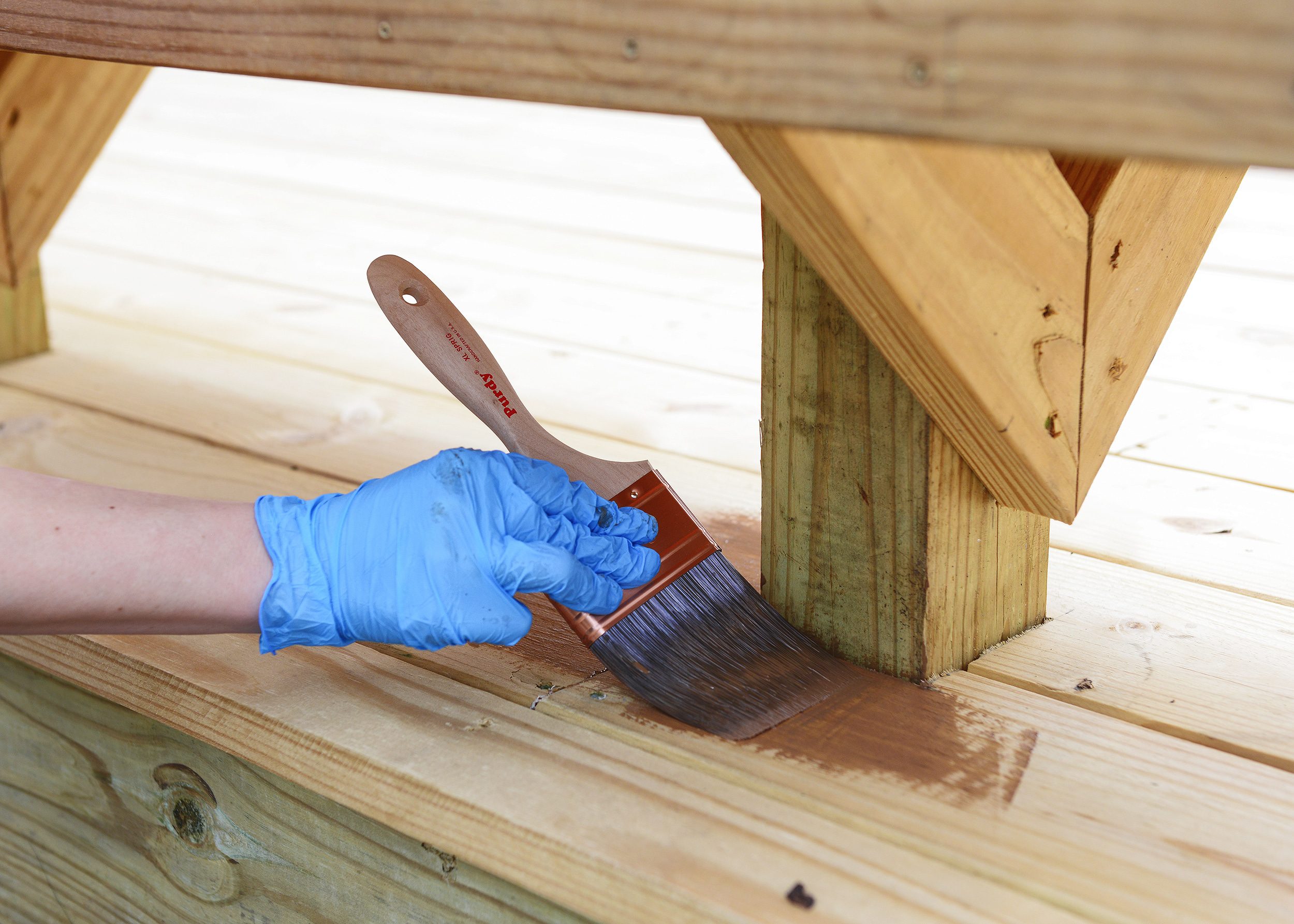 A woman's hand in a blue glove applies stain under a deck bench // via Yellow Brick Home