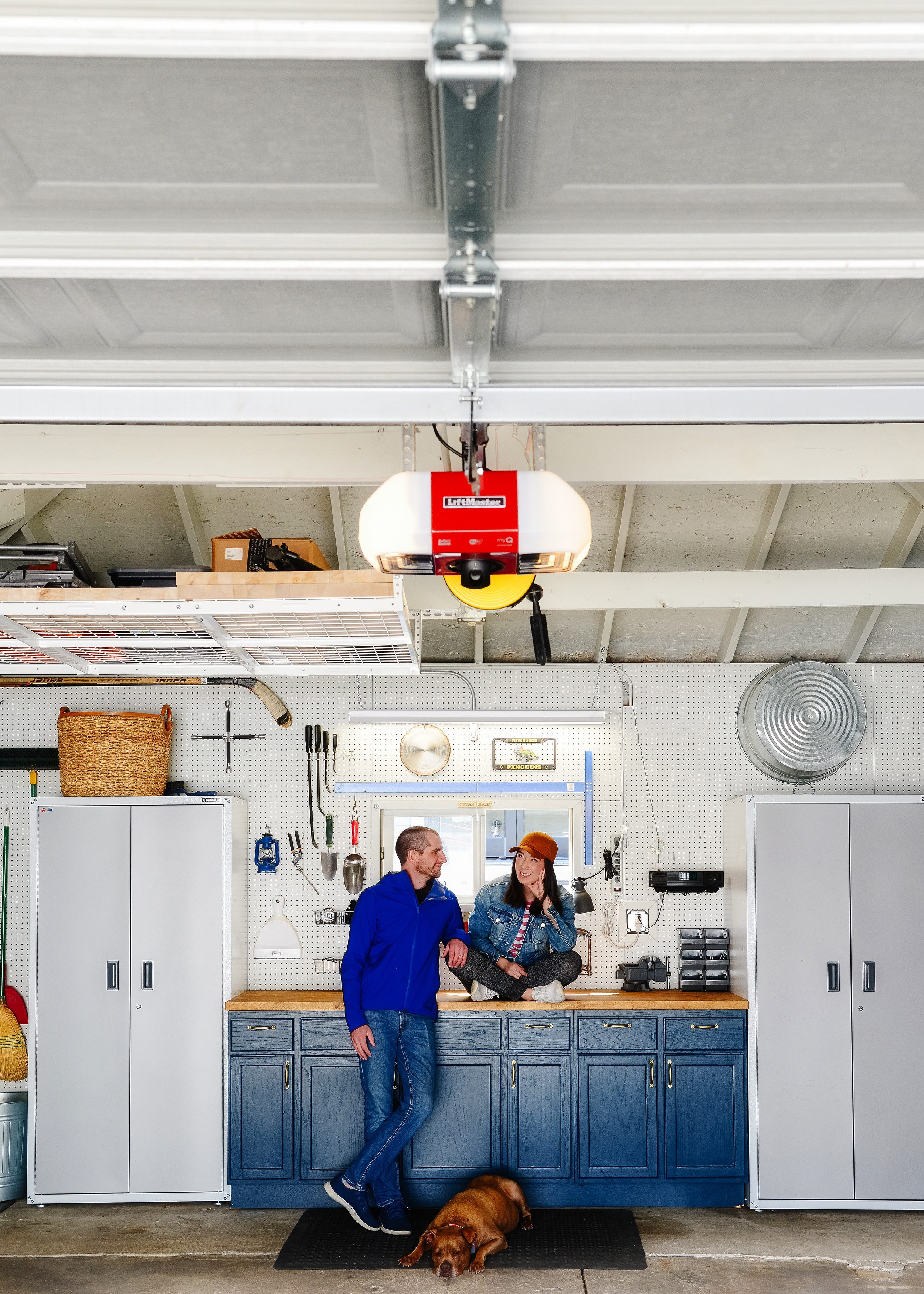 A husband and wife sit on a blue workbench in a tidy garage with a new red Liftmaster garage door opener // via yellow brick home  