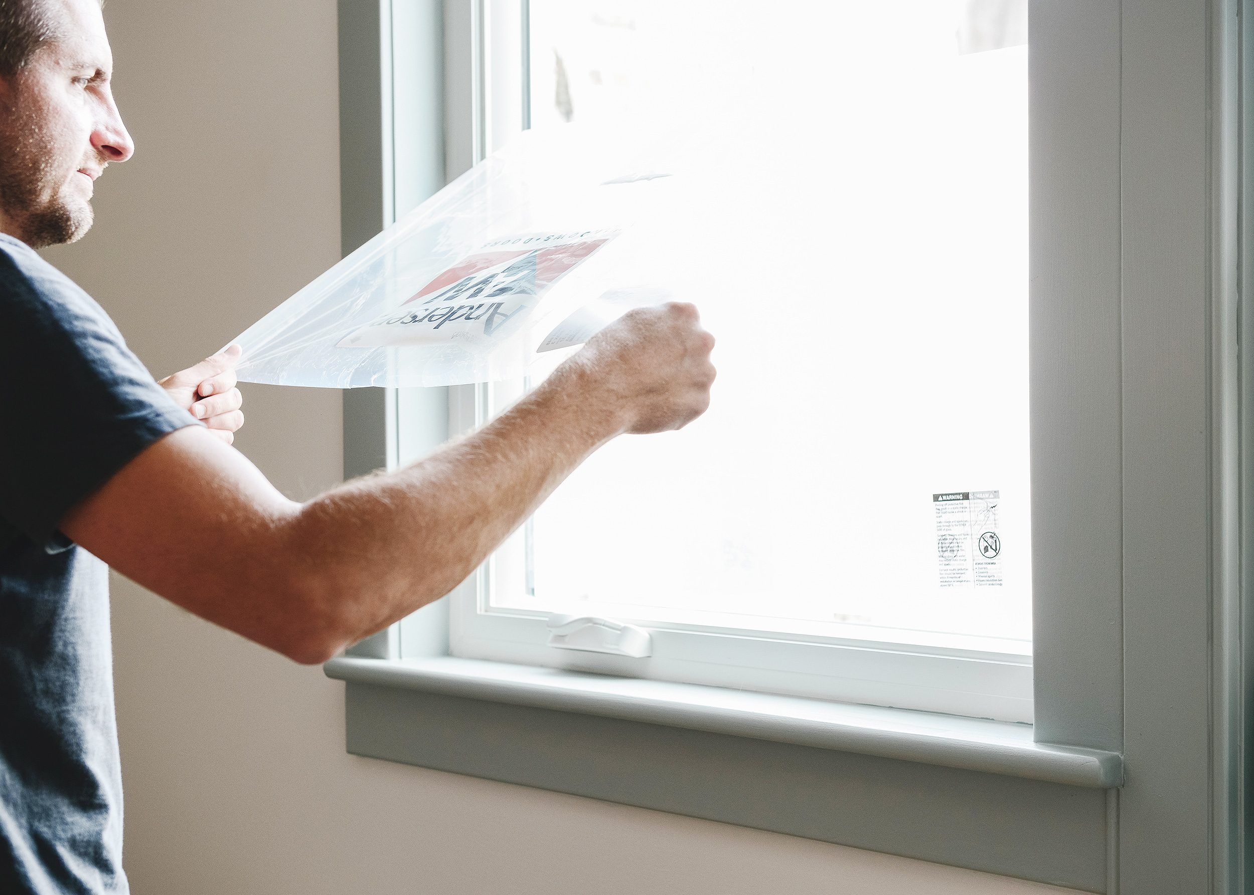 A man pulls protective window film  from a newly installed Andersen window in a white room with grey-blue contrasting trim // via Yellow Brick Home