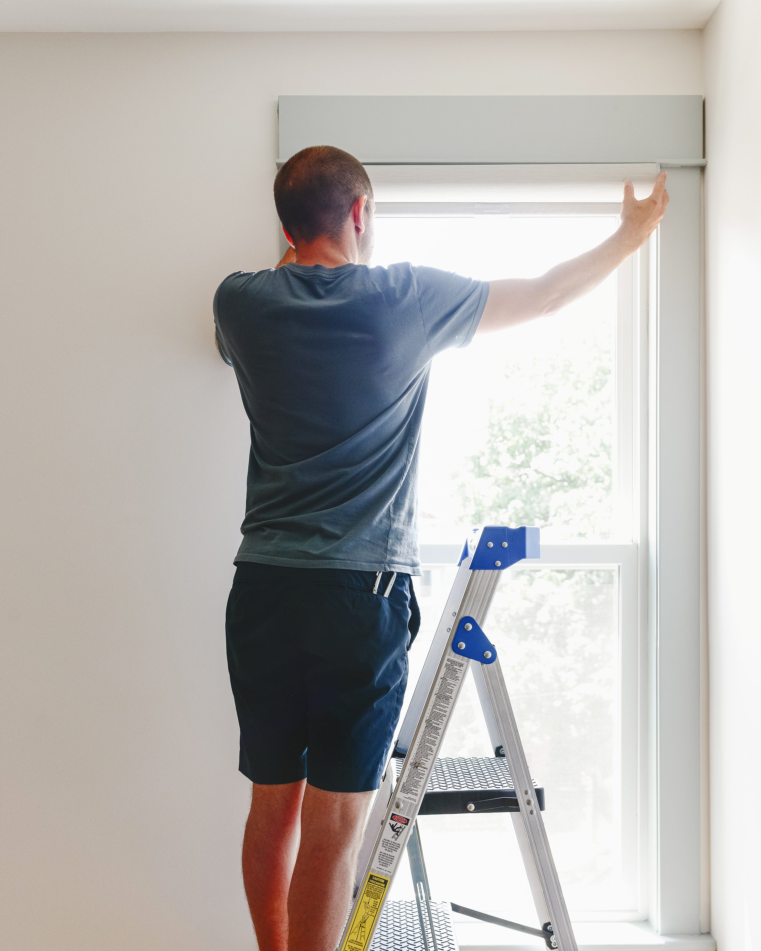 A man on a ladder installs a roller shade mounting bracket inside a window // via Yellow Brick Home