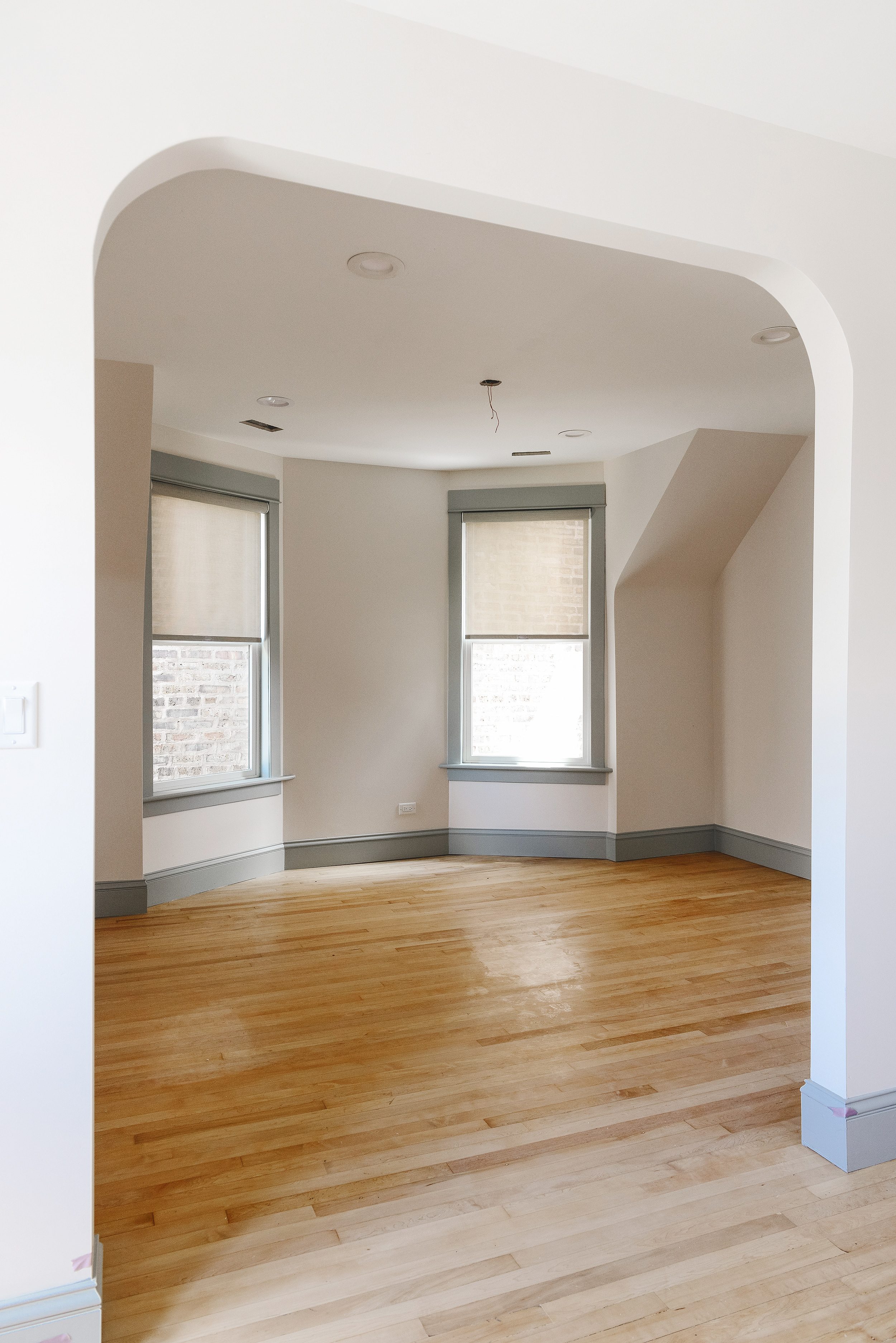 An arched doorway opening  in a renovated Chicago apartment features Bali blinds installed in large windows with Sherwin Williams Magnetic Gray trim on Sherwin Williams Heron Plume walls atop refinished vintage maple floors // via Yellow Brick Home