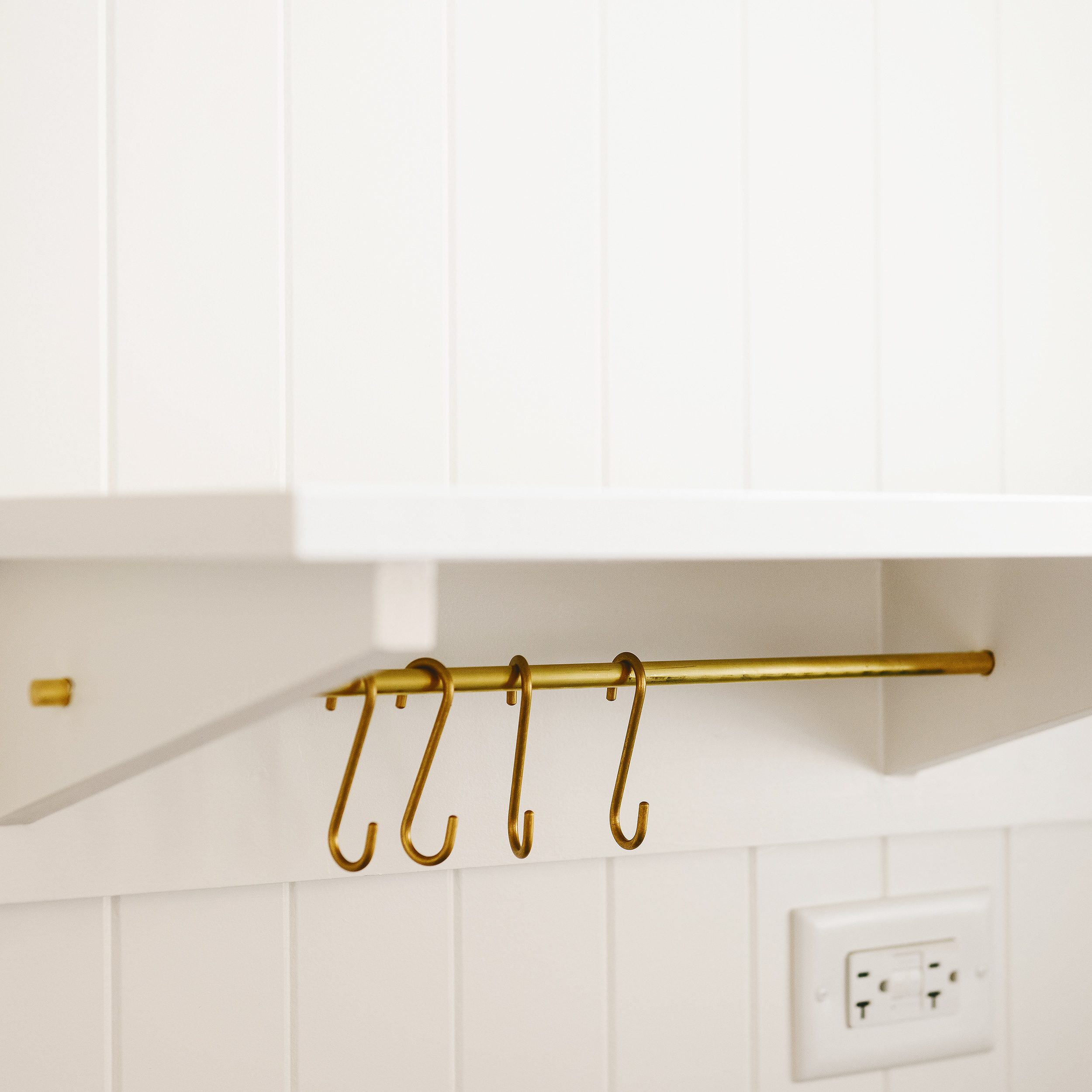 A custom white floating shelf with an integrated brass hanging rod with four brass 'S' hooks. 