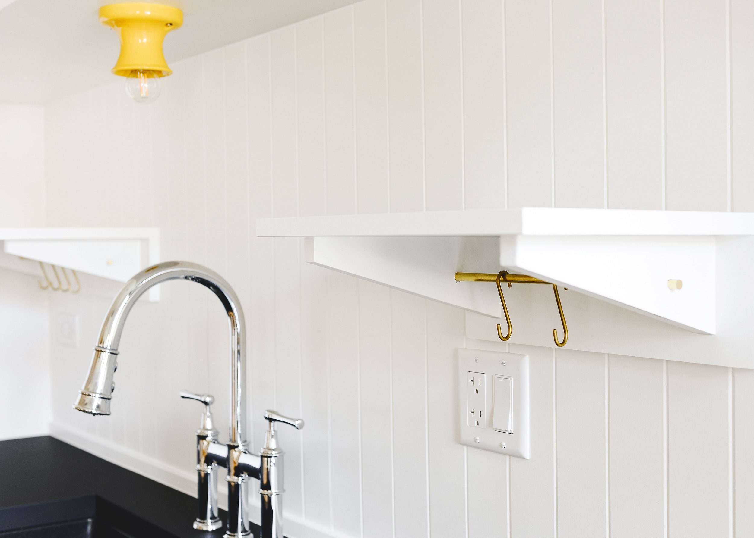 A pair of custom white floating shelves with integrated brass rods and 'S' hooks are flanked by a chrome bridge faucet below and a yellow ceramic sconce above