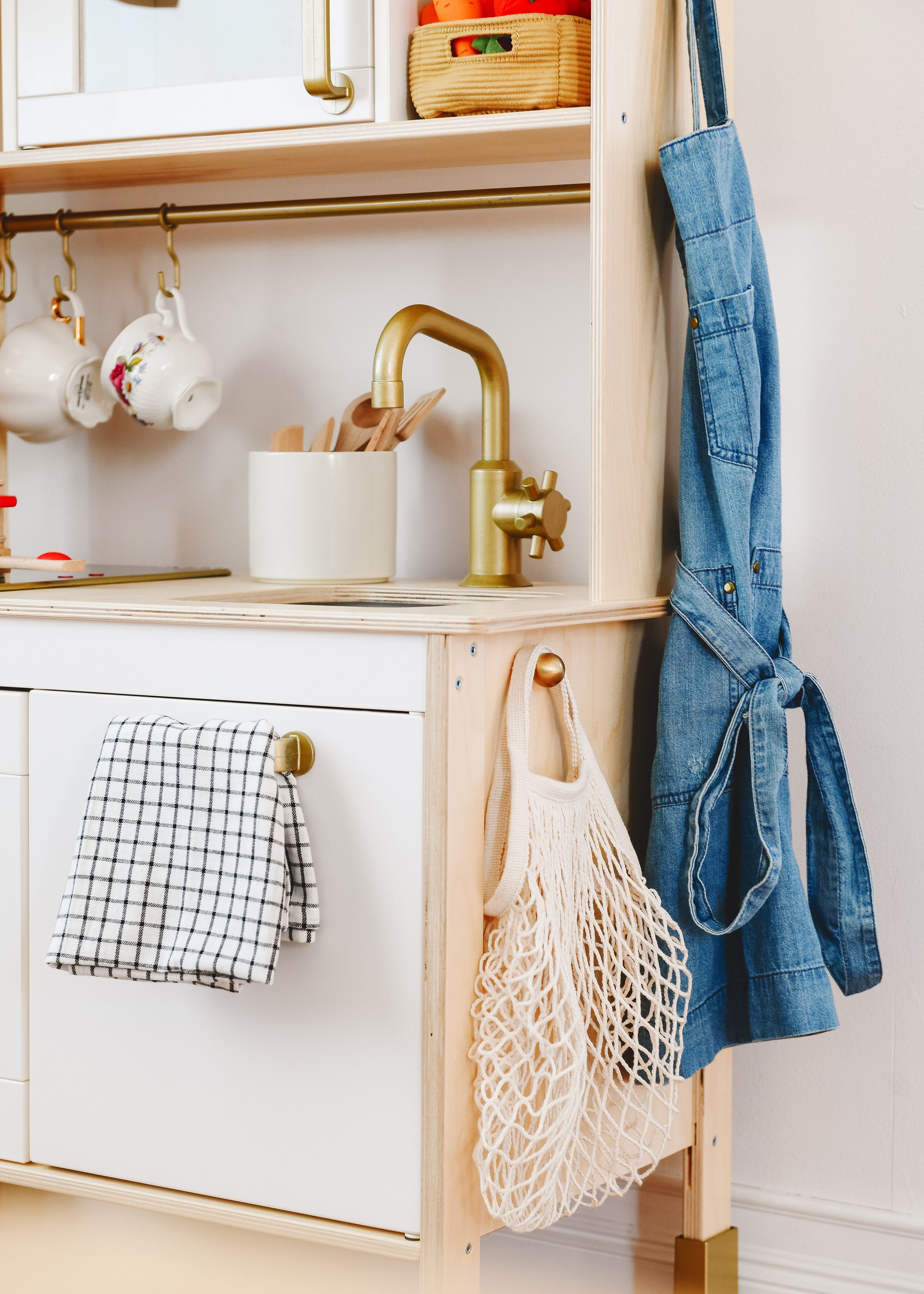 Tips for Stocking Your Play Kitchen + 36 of the Cutest Accessories! | A corner of Lucy's playroom showing a close up of her IKEA DUKTIG play kitchen and hanging brass knobs for her apron and mesh bag | via Yellow Brick Home
